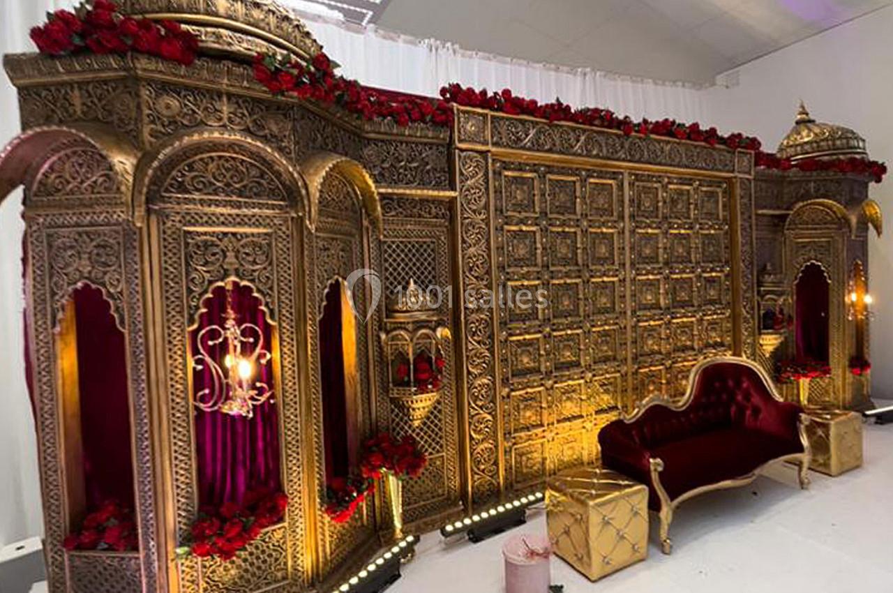 Décor de mariage avec une structure dorée ornée de motifs complexes, roses rouges et canapé en velours bordeaux.