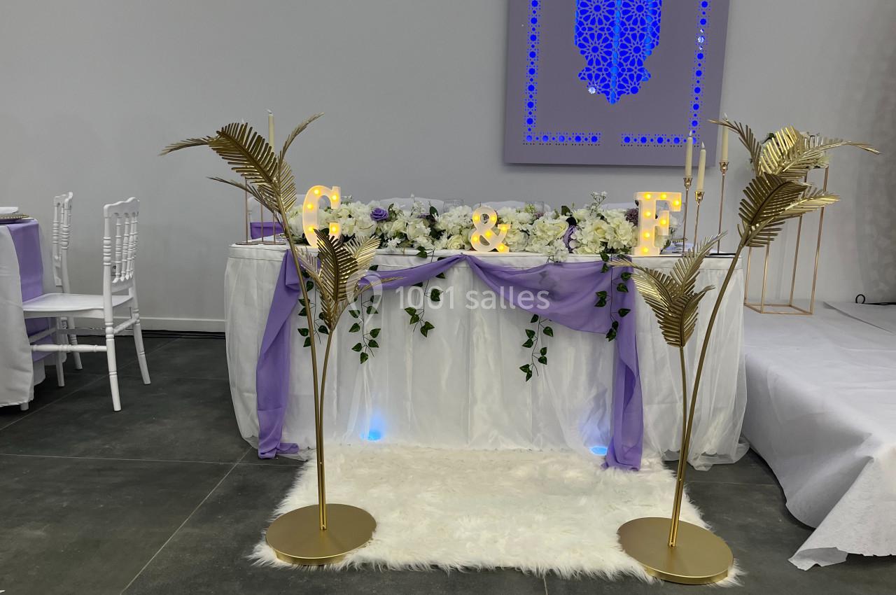 Décoration de table de mariage avec fleurs blanches, tissu violet, lettres lumineuses et ornements dorés.