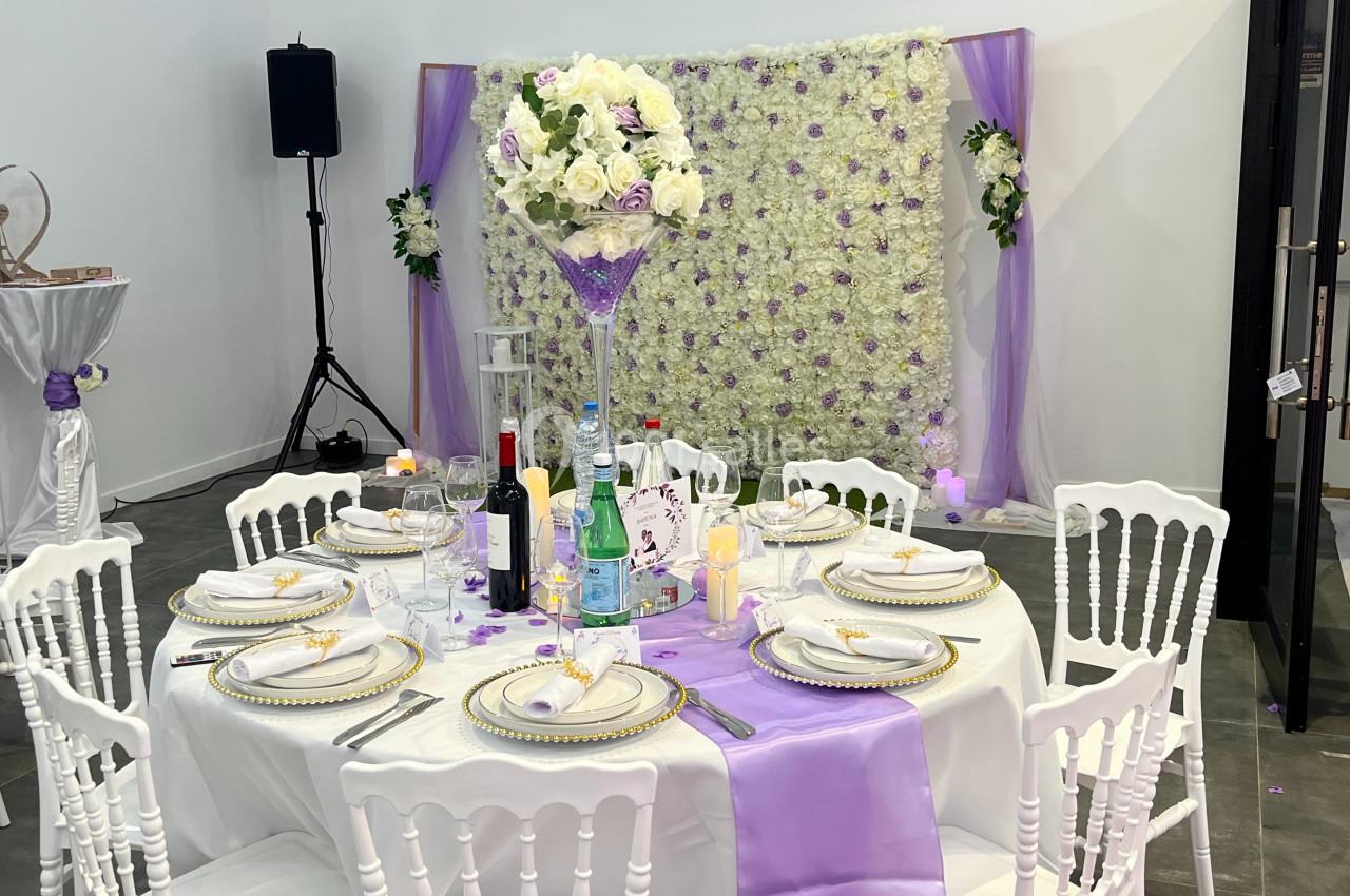 Table décorée pour un événement avec nappes blanches, accents violets, vaisselle élégante et un mur floral en arrière-plan.