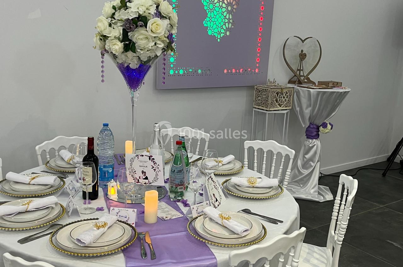 Table décorée pour un événement avec nappes blanches, chemin de table violet, vaisselle élégante et centre de table floral.