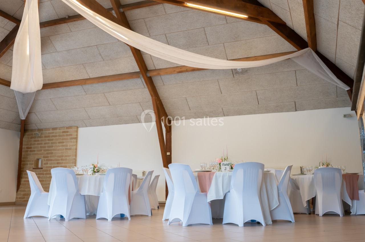 Salle de réception décorée avec des tables rondes, nappes blanches et chaises couvertes, sous un plafond en bois.