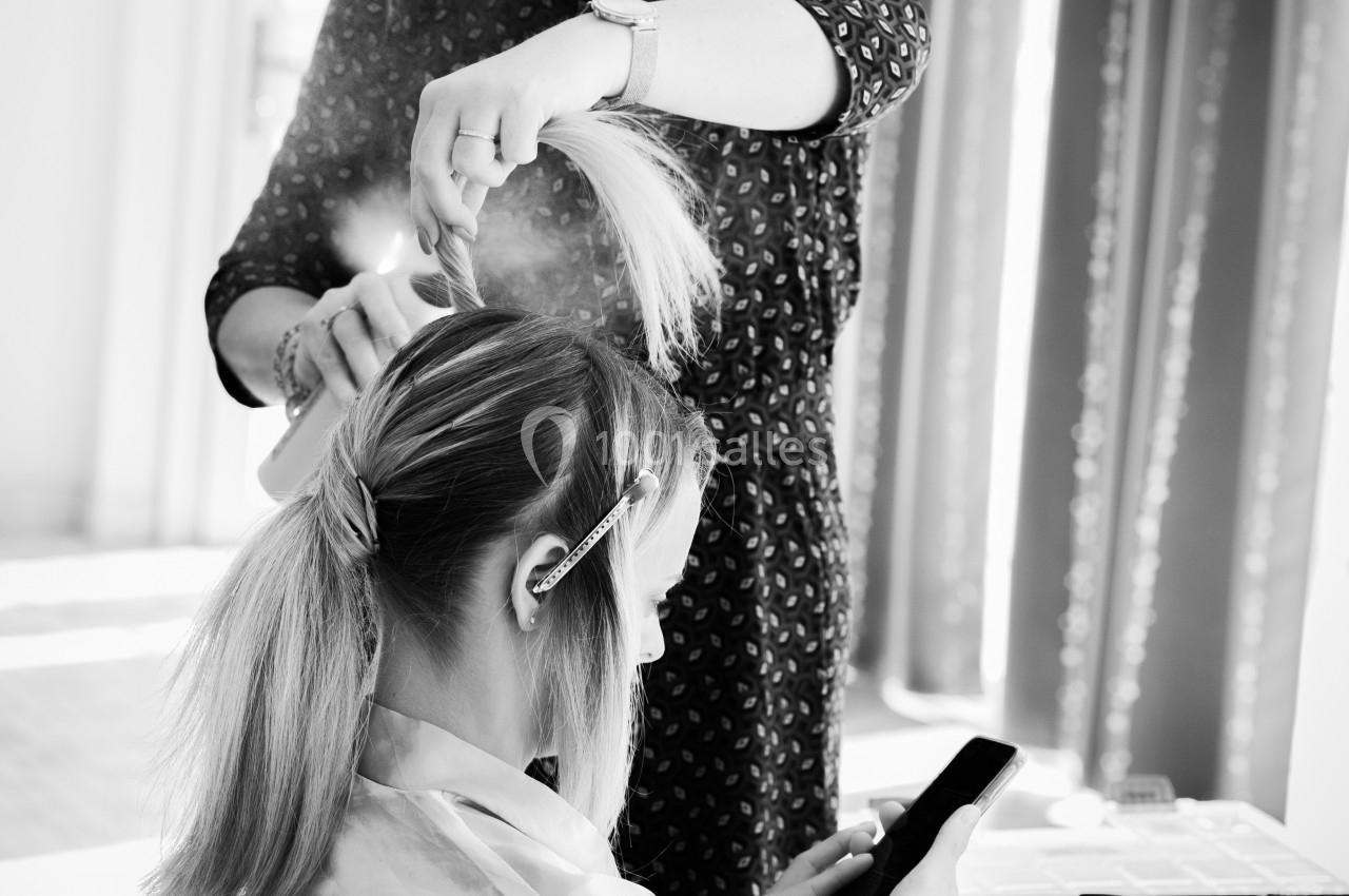 Une coiffeuse attache les cheveux d'une cliente assise, concentrée sur son téléphone, dans un salon lumineux.