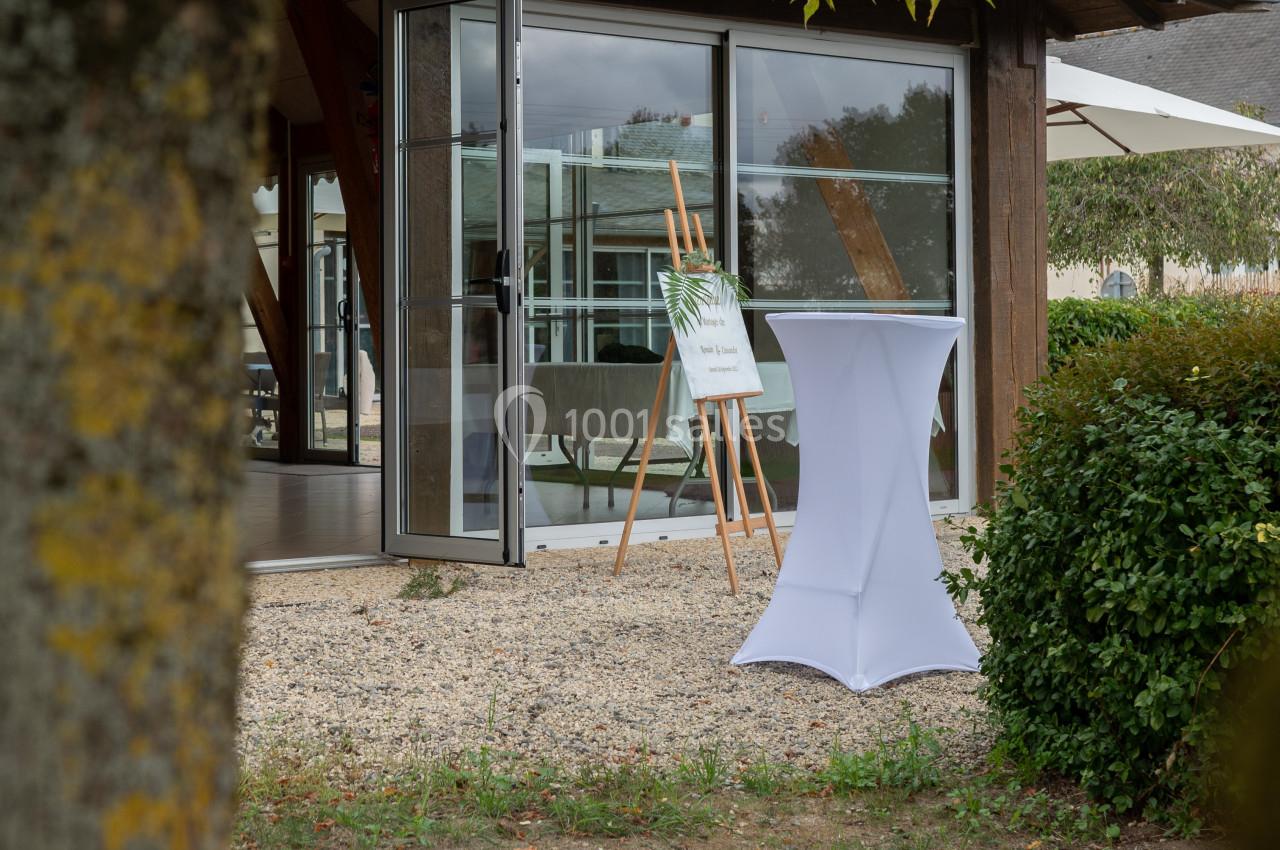 Entrée d'un bâtiment avec porte vitrée ouverte, table haute blanche et chevalet sur une allée gravillonnée.