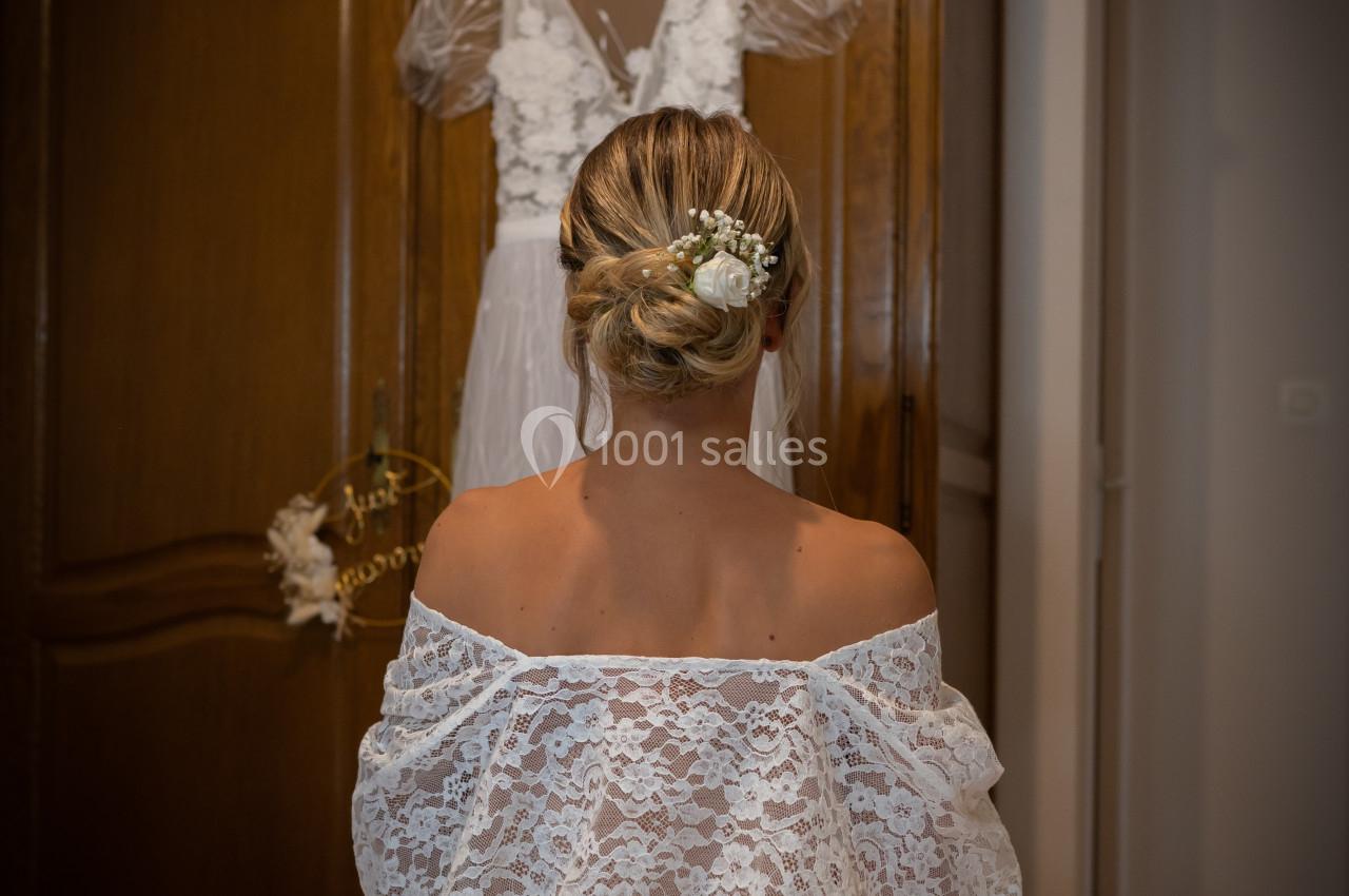 Une femme en robe en dentelle blanche, de dos, assise face à une armoire où est suspendue une robe de mariée.