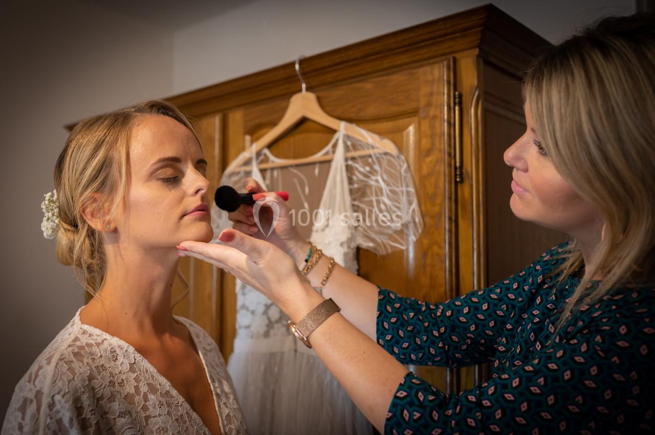 Une maquilleuse applique du maquillage sur le visage d'une femme en robe blanche devant une armoire en bois.