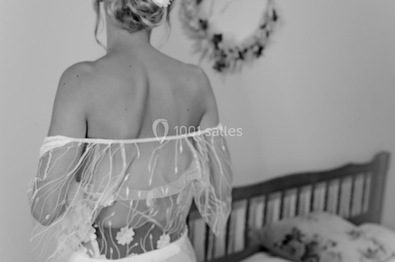 Femme de dos en robe de mariée avec un chignon décoré, debout près d'un lit dans une pièce lumineuse.