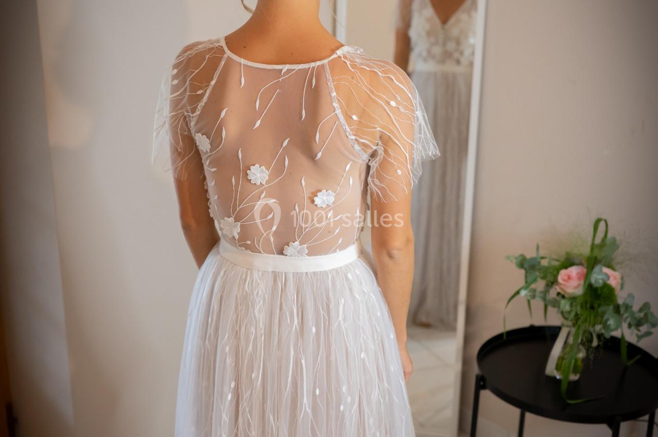 Une femme en robe de mariée vue de dos, se tenant devant un miroir dans une pièce lumineuse.