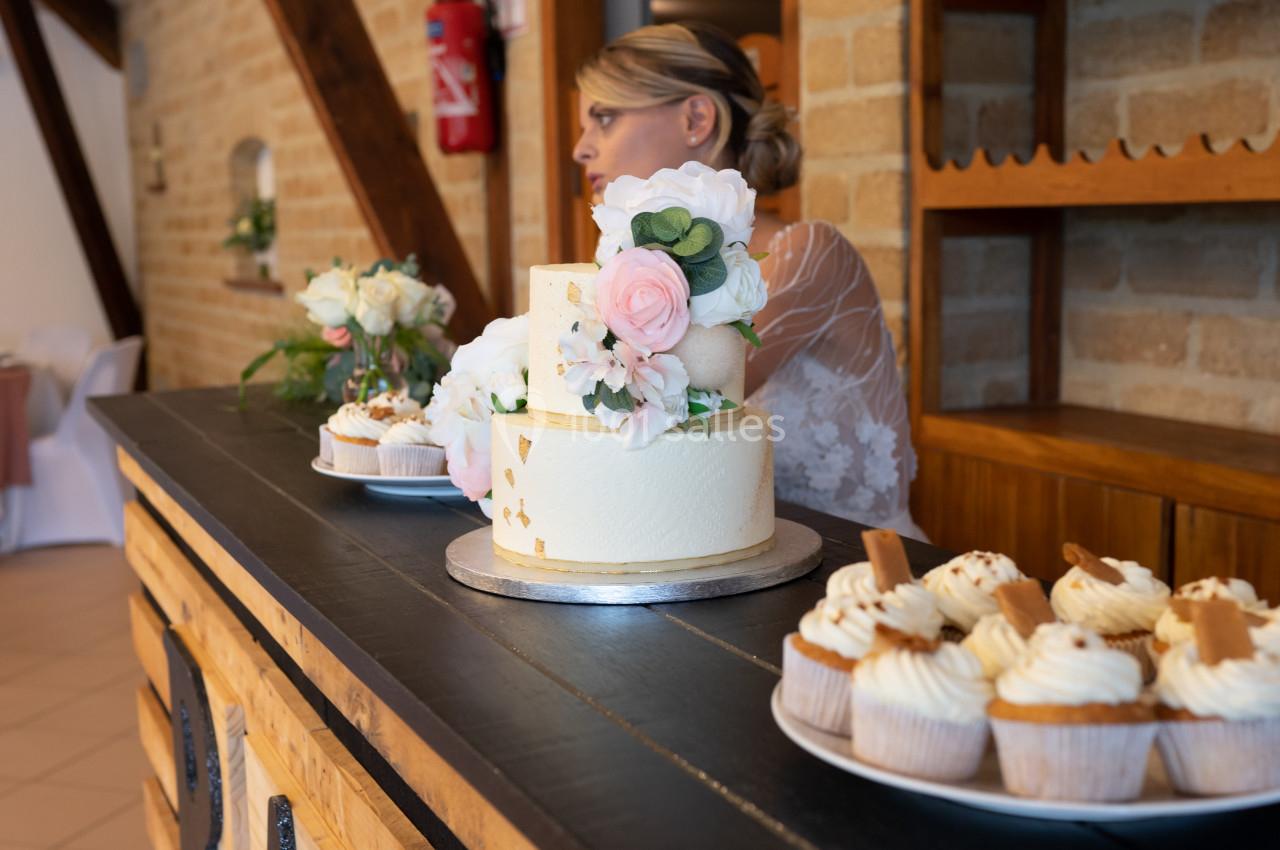 Gâteau de mariage décoré de fleurs et cupcakes sur un comptoir en bois, avec une femme en arrière-plan.