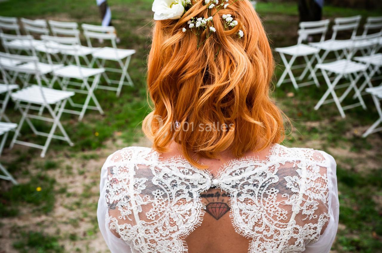 Une mariée rousse de dos, portant une robe en dentelle, face à des chaises blanches disposées en extérieur.