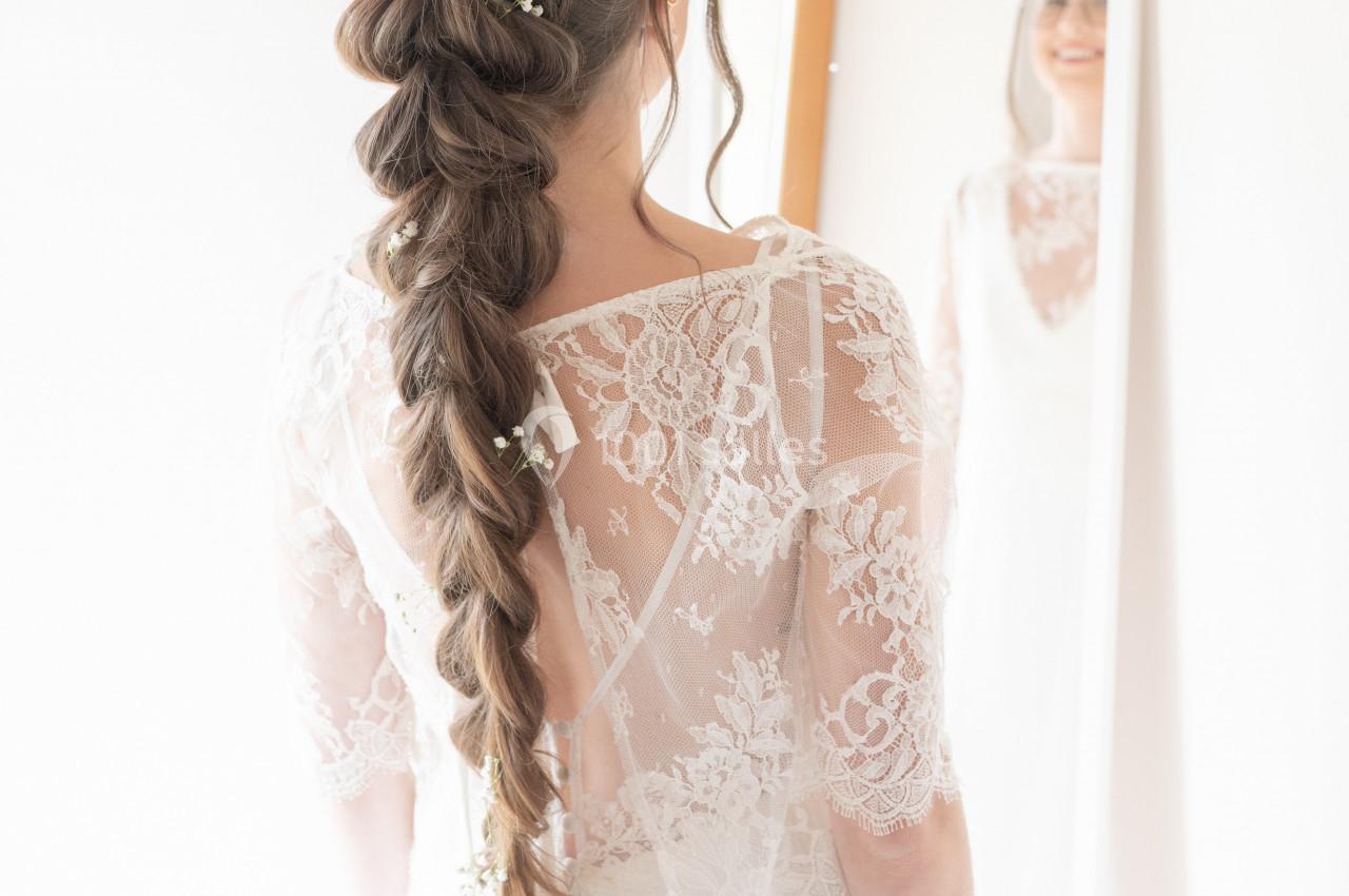 Femme en robe blanche avec une longue tresse ornée de fleurs, se regardant dans un miroir.