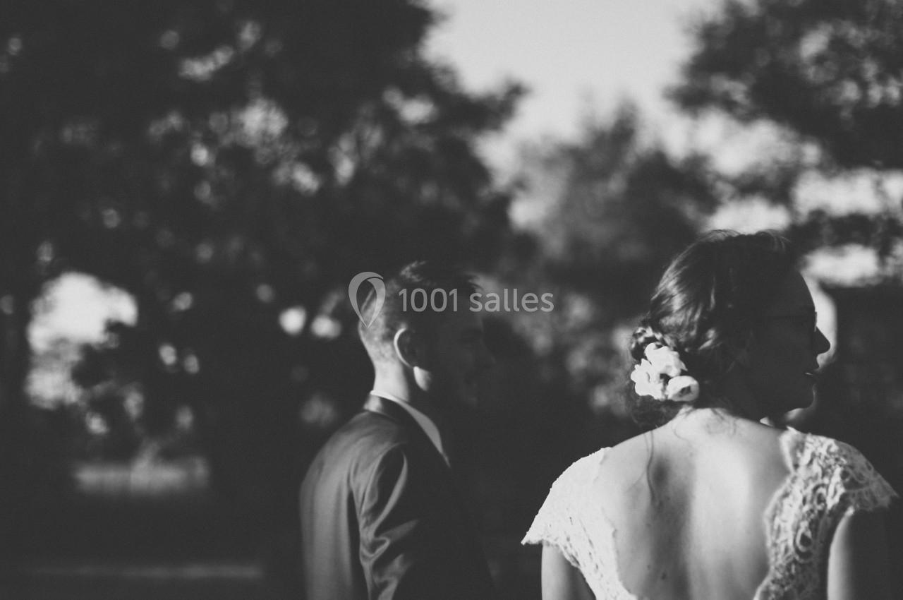 Un couple en tenue de mariage, vu de dos, se tenant la main dans un cadre extérieur en noir et blanc.