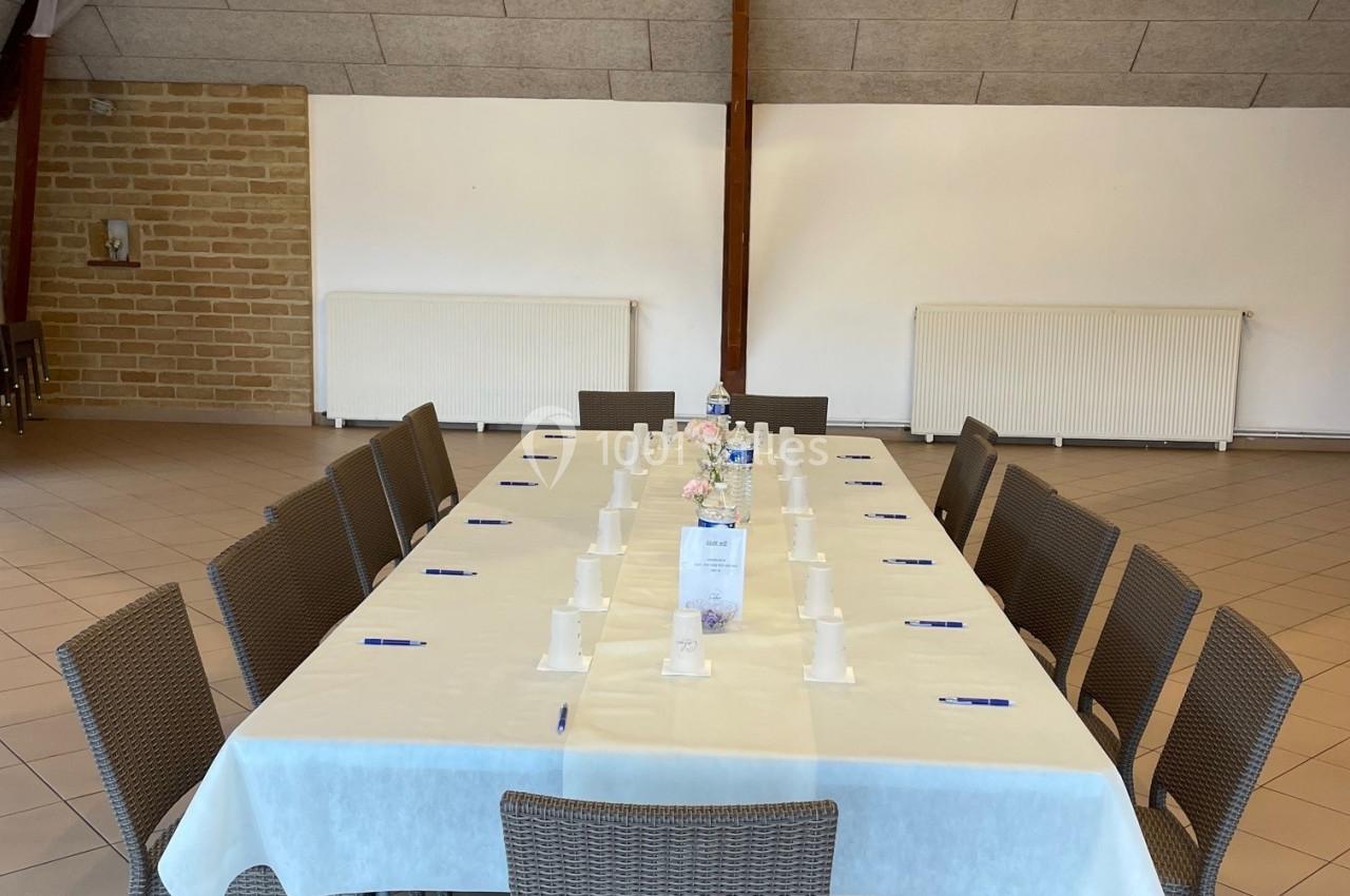 Table rectangulaire dressée avec nappes blanches, chaises en osier, stylos et gobelets, dans une salle au plafond en bois.