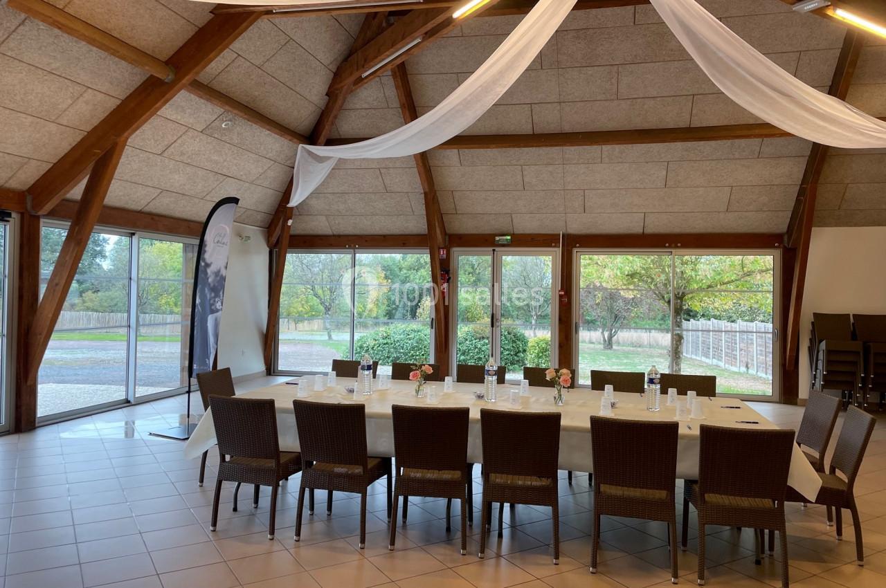 Salle lumineuse avec grande table rectangulaire dressée, chaises en rotin, baies vitrées donnant sur un jardin.