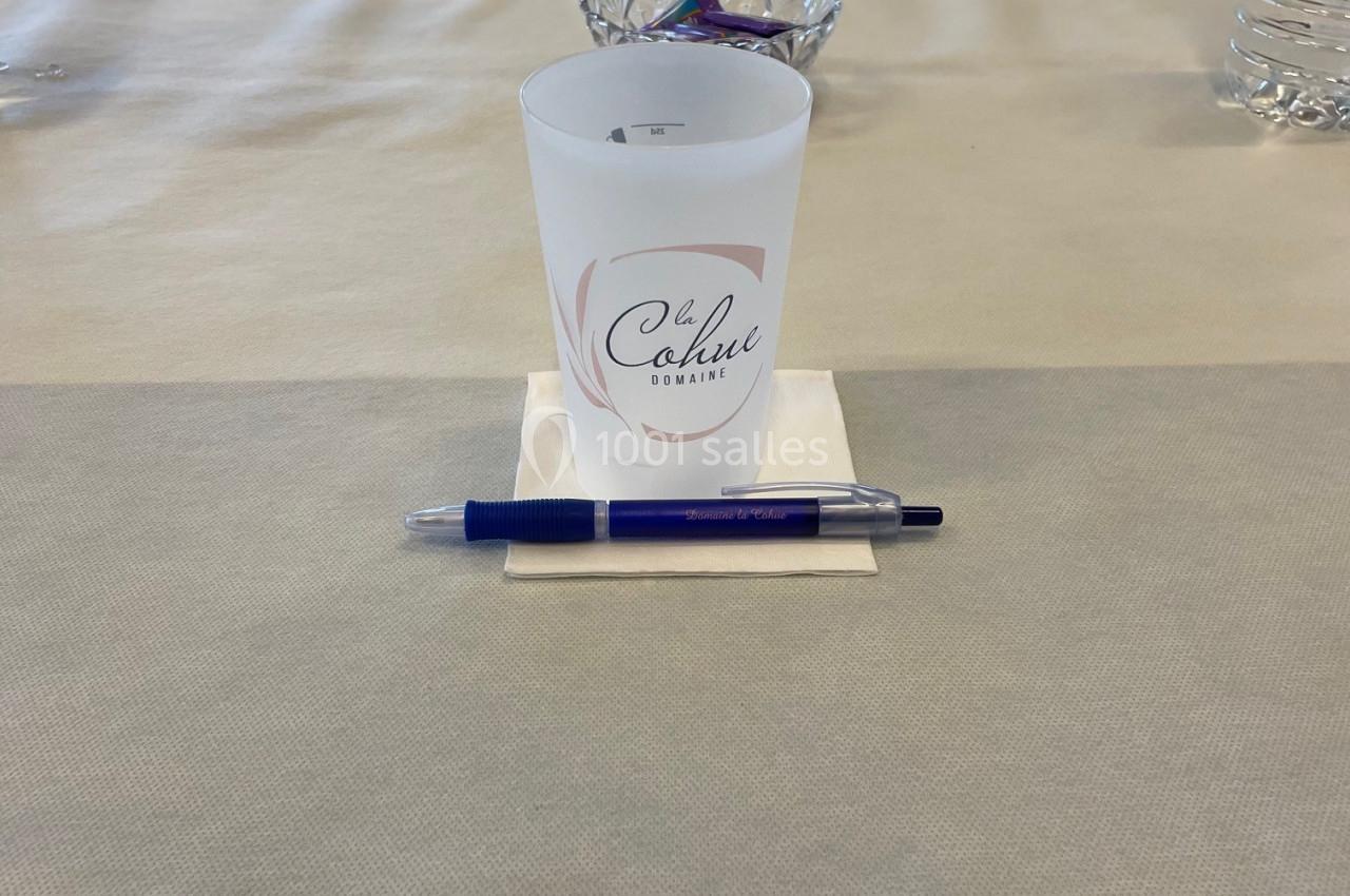 Un gobelet personnalisé, un stylo bleu et une bouteille d'eau disposés sur une table beige dans une salle lumineuse.