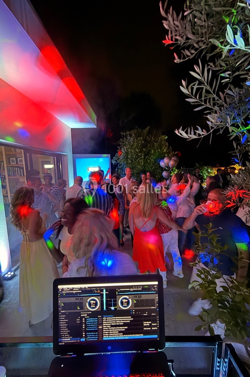 Groupe de personnes dansant en soirée en extérieur, éclairées par des lumières colorées, avec une table de mixage au premier…