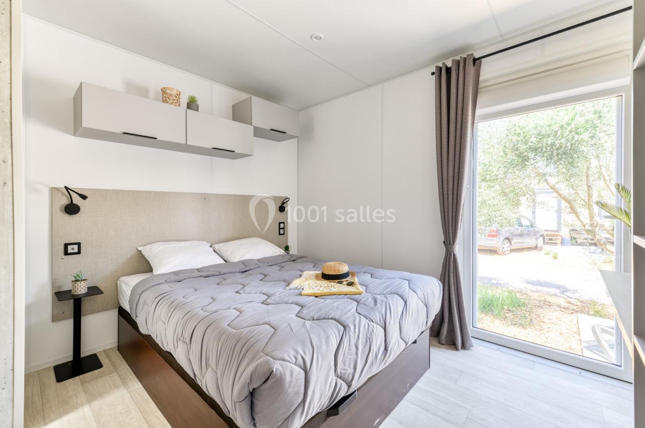 Chambre lumineuse avec lit double, couvre-lit gris, étagères murales, rideaux gris et vue sur un extérieur arboré.