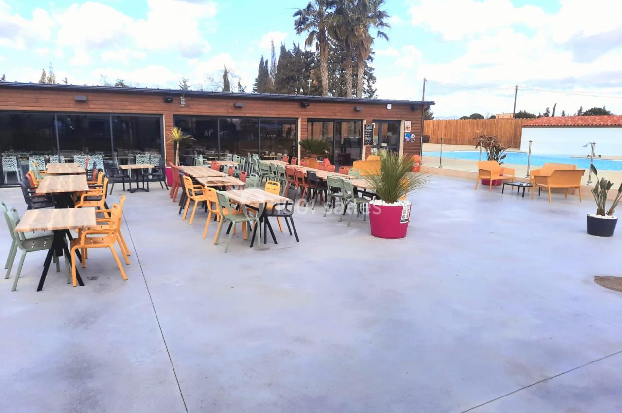 Terrasse extérieure avec tables et chaises colorées, bordée de plantes en pots, proche d'une piscine et d'un bâtiment en…