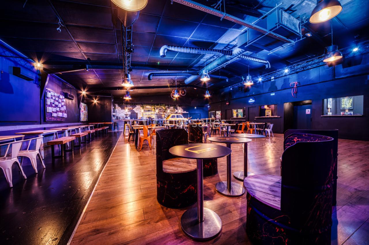 Salle moderne avec éclairage tamisé, tables en bois, chaises variées et décoration industrielle.