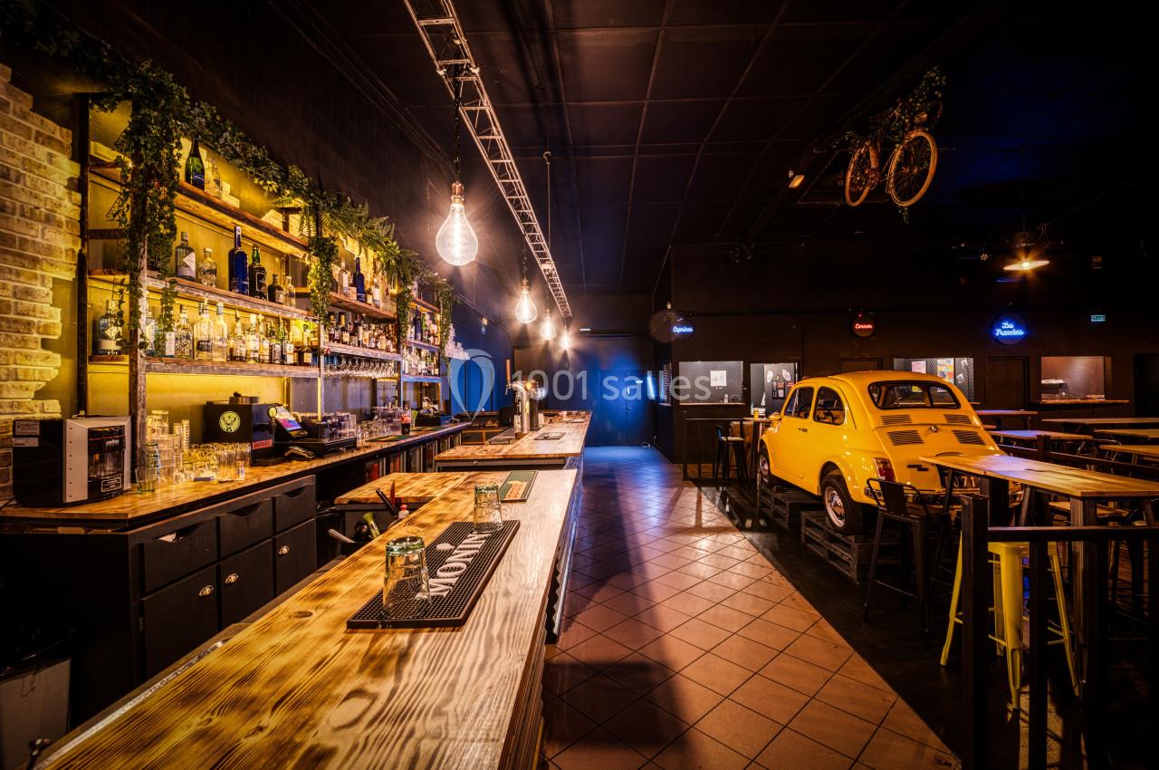 Intérieur d'un bar avec un long comptoir en bois, étagères garnies de bouteilles et une voiture jaune en décoration.