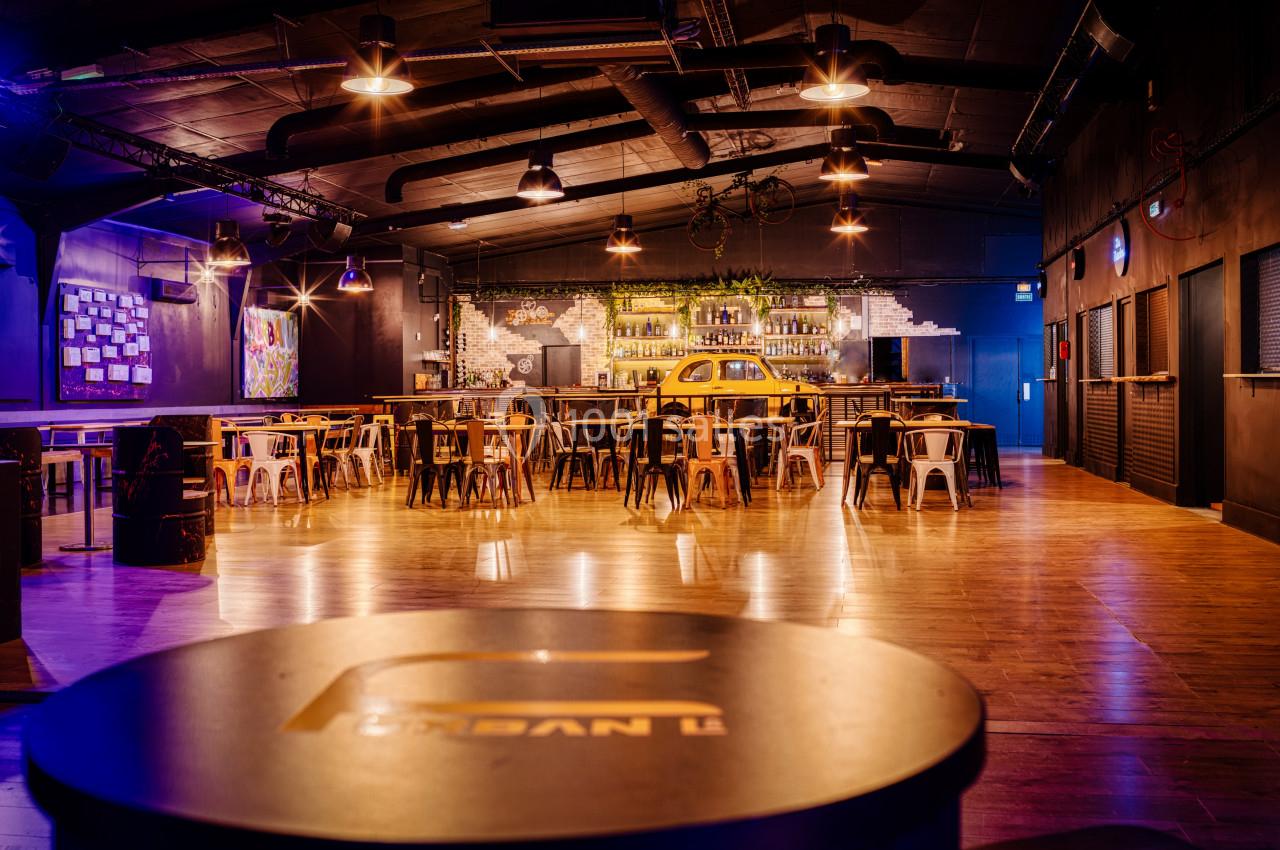 Salle spacieuse avec tables, chaises, bar éclairé et décoration industrielle, ambiance chaleureuse et moderne.