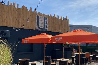 Terrasse extérieure avec tables, chaises et parasols orange, entourée de végétation, devant un bâtiment en bois et métal.