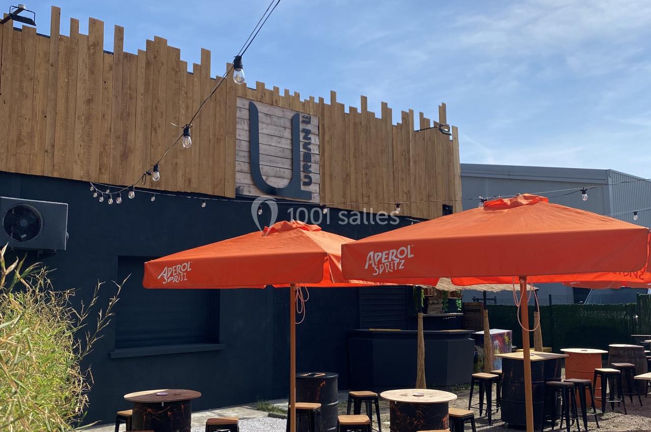 Terrasse extérieure avec tables, chaises et parasols orange, entourée de végétation, devant un bâtiment en bois et métal.