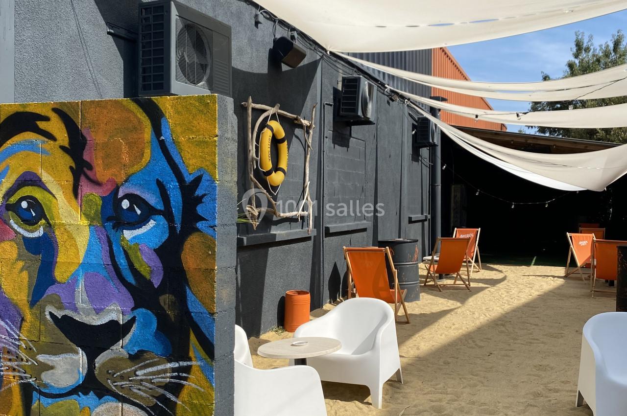 Mur peint avec un lion coloré, terrasse extérieure avec chaises longues sur du sable sous des voiles d'ombrage.