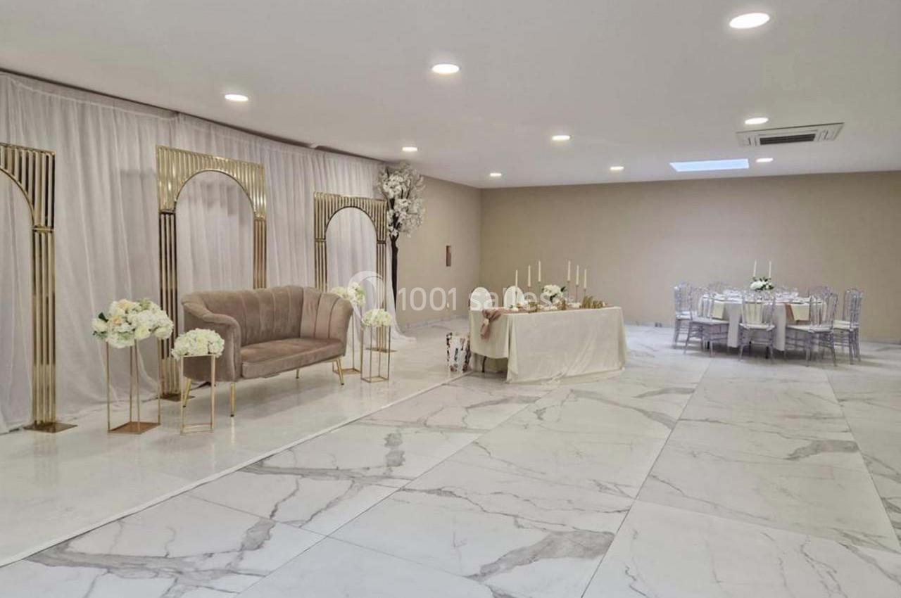 Salle de réception lumineuse avec sol en marbre, arche dorée, canapé beige et tables décorées pour un événement.