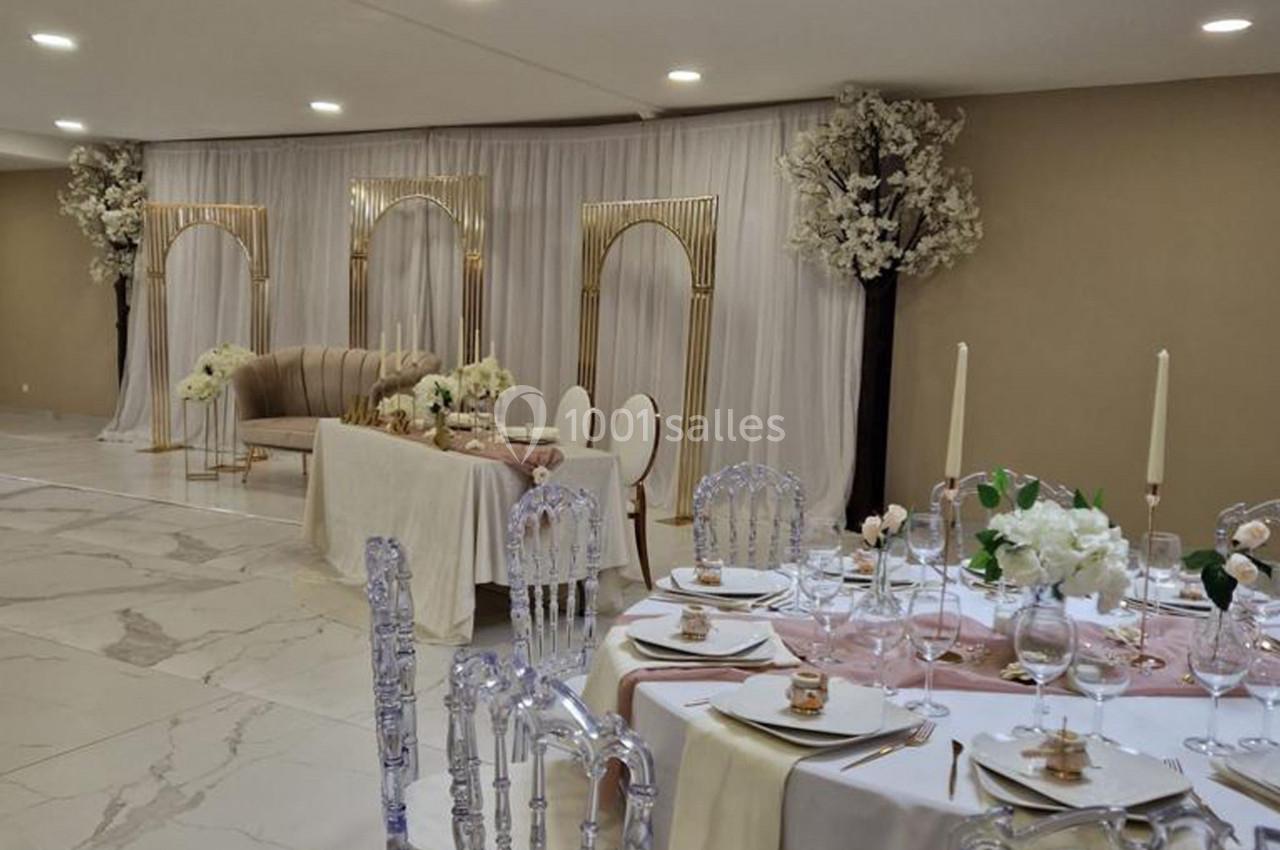 Salle de réception élégante avec tables dressées, chaises transparentes et décorations florales blanches et dorées.