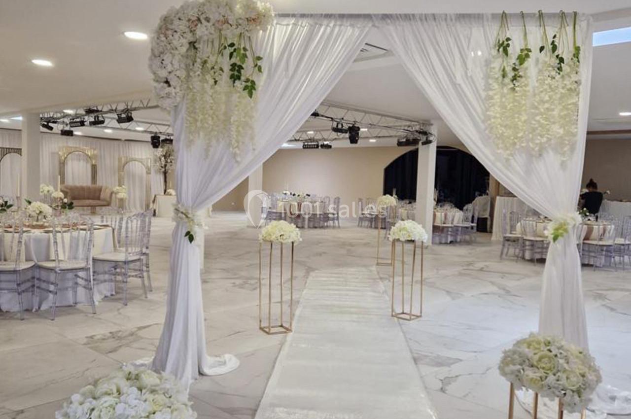 Allée décorée pour une cérémonie, avec arche florale blanche, chaises transparentes et tables dressées en arrière-plan.