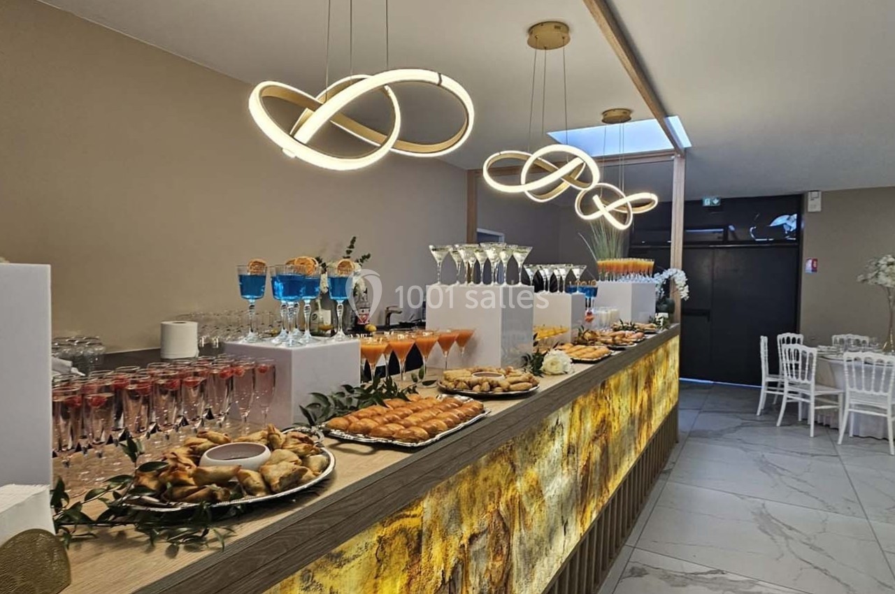 Buffet élégant avec boissons, verrines et amuse-bouches, dans une salle lumineuse avec tables dressées en arrière-plan.