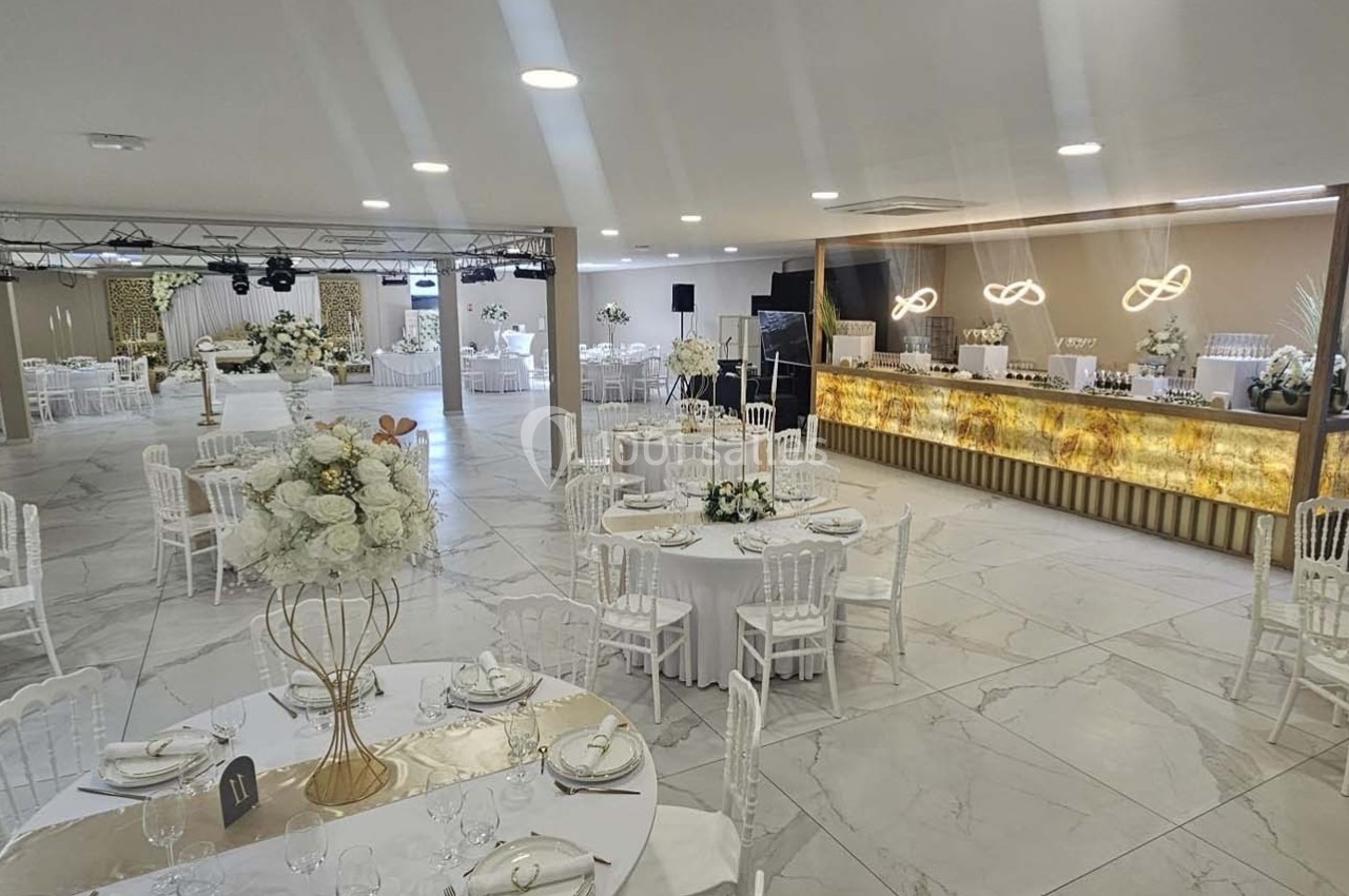 Salle de réception élégante avec tables rondes décorées, chaises blanches et un bar éclairé en arrière-plan.