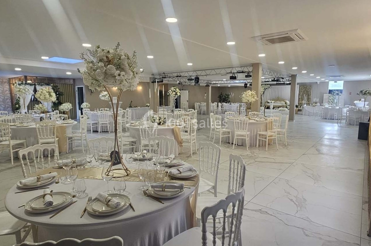 Salle de réception élégamment décorée avec tables rondes dressées, chaises blanches et arrangements floraux.