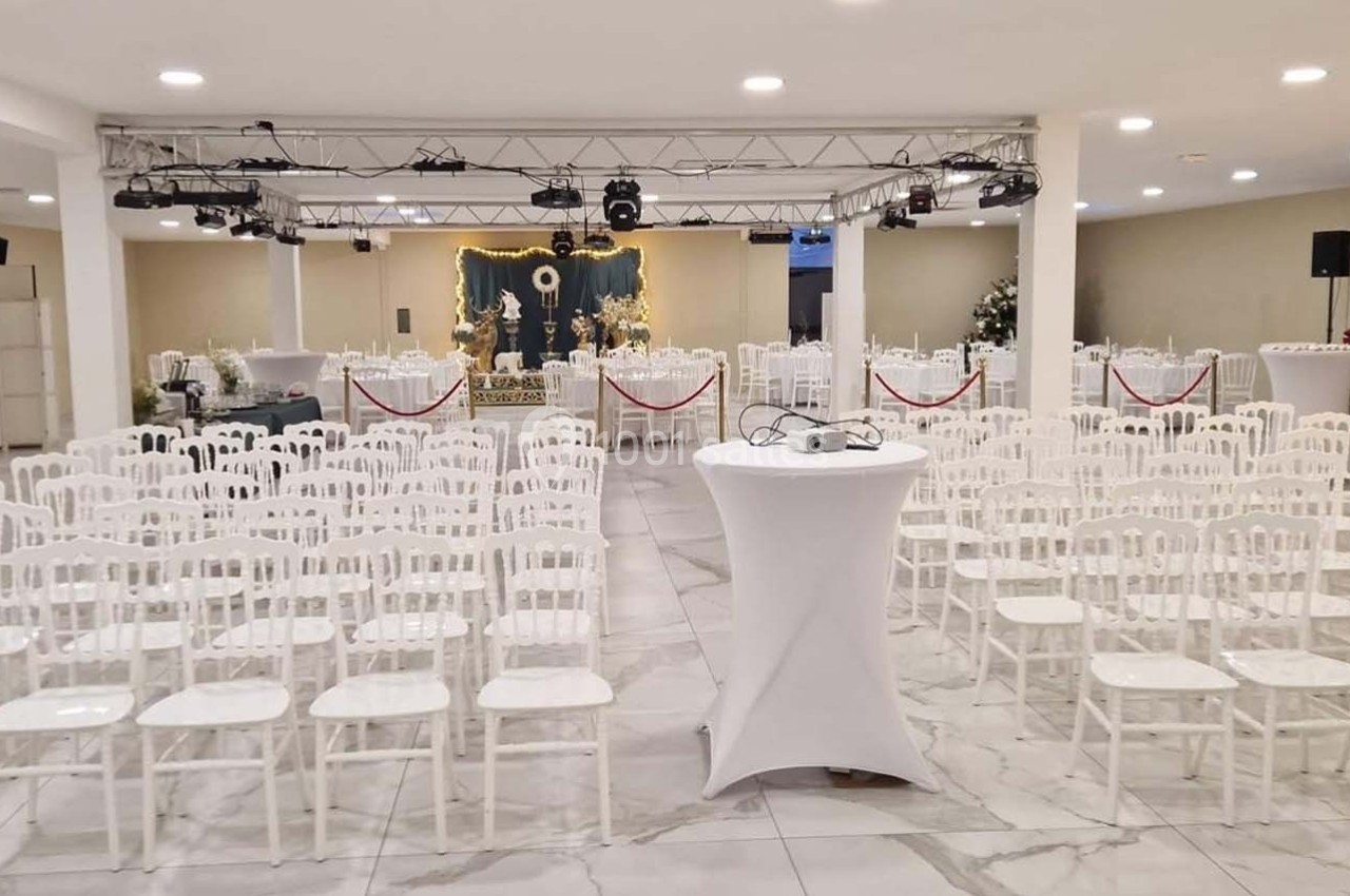 Salle de réception avec rangées de chaises blanches alignées, une scène décorée et des éclairages suspendus.