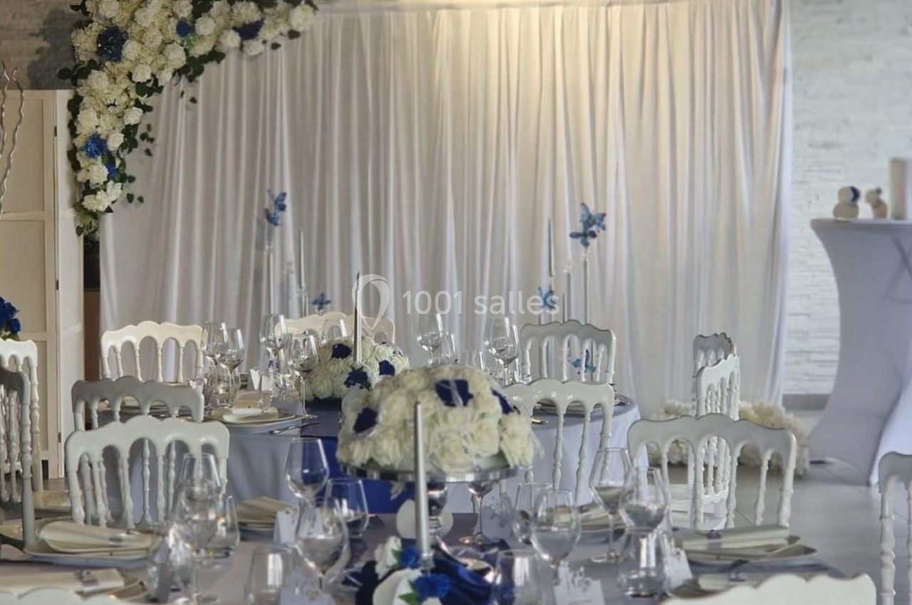 Salle décorée pour un événement avec des tables dressées, des chaises blanches et des arrangements floraux bleus et blancs.