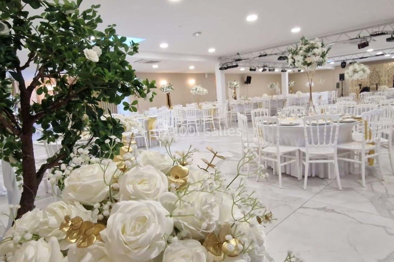 Salle de réception élégante avec tables rondes dressées, chaises blanches et décorations florales blanches et dorées.