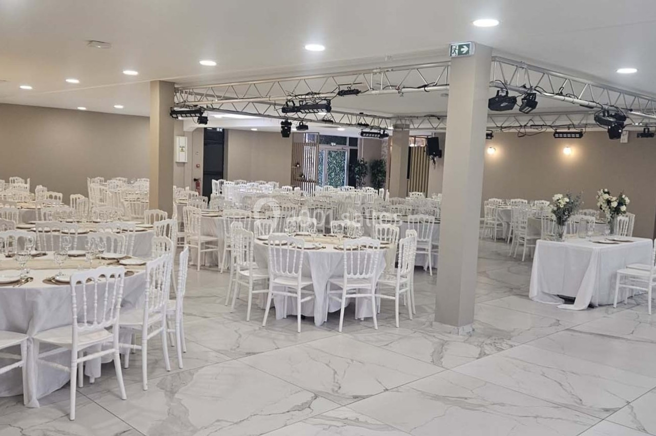 Salle de réception lumineuse avec tables rondes dressées, chaises blanches et décorations florales.