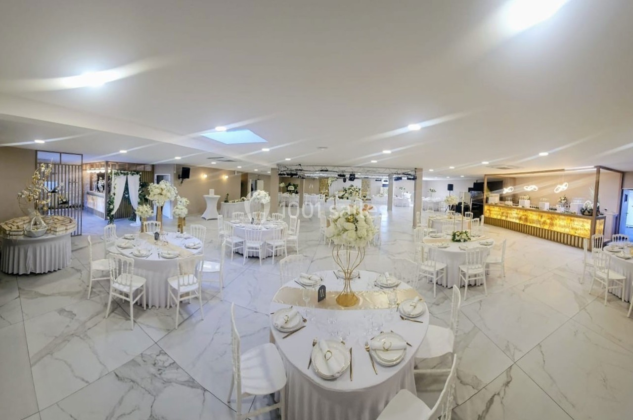 Salle de réception élégante avec tables rondes dressées, décorations florales blanches et éclairage lumineux.