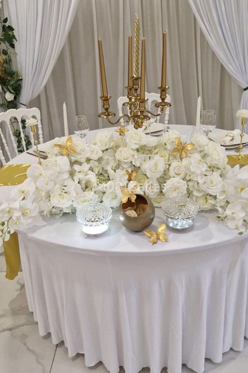 Table décorée avec des fleurs blanches, des chandeliers dorés et des papillons dorés, sur fond de rideaux clairs.