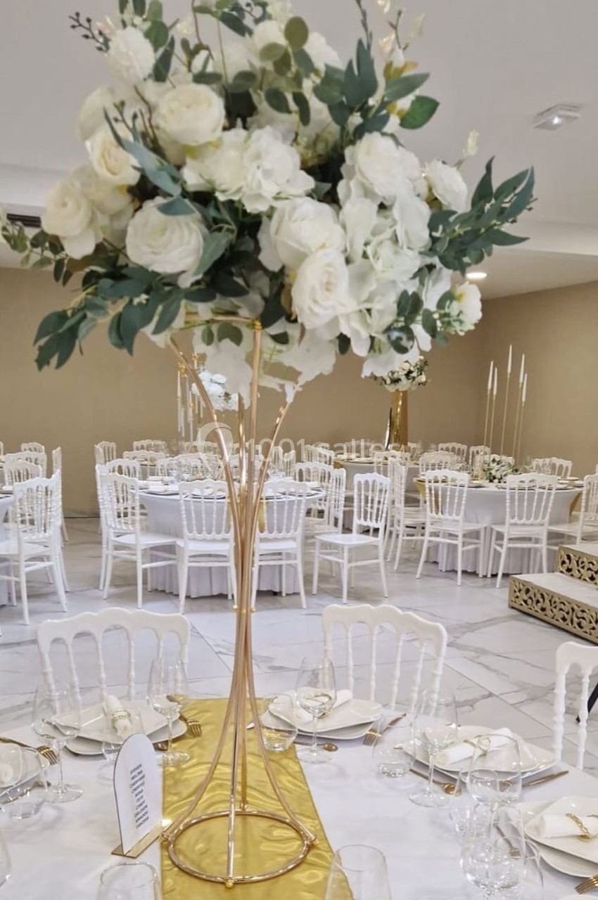 Centre de table floral avec roses blanches et feuillage sur support doré, entouré de tables dressées avec chaises blanches.