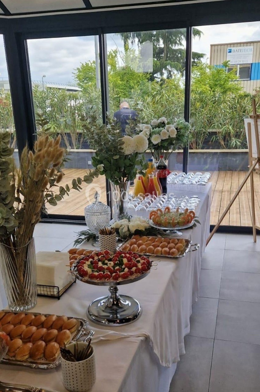 Buffet dressé avec viennoiseries, fruits, jus et décorations florales, près de baies vitrées donnant sur une terrasse.