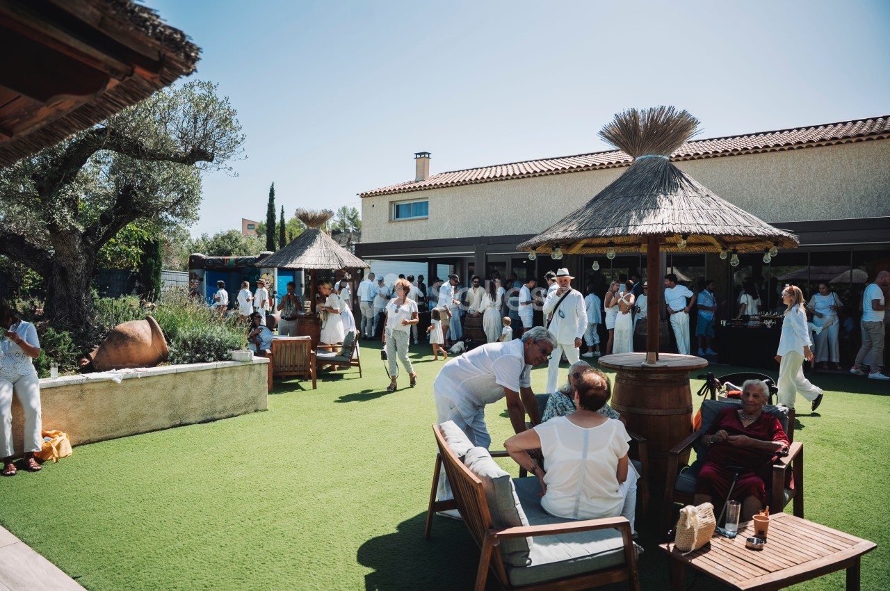 Personnes rassemblées dans un jardin ensoleillé avec des parasols en paille, discutant et profitant d'un moment convivial.