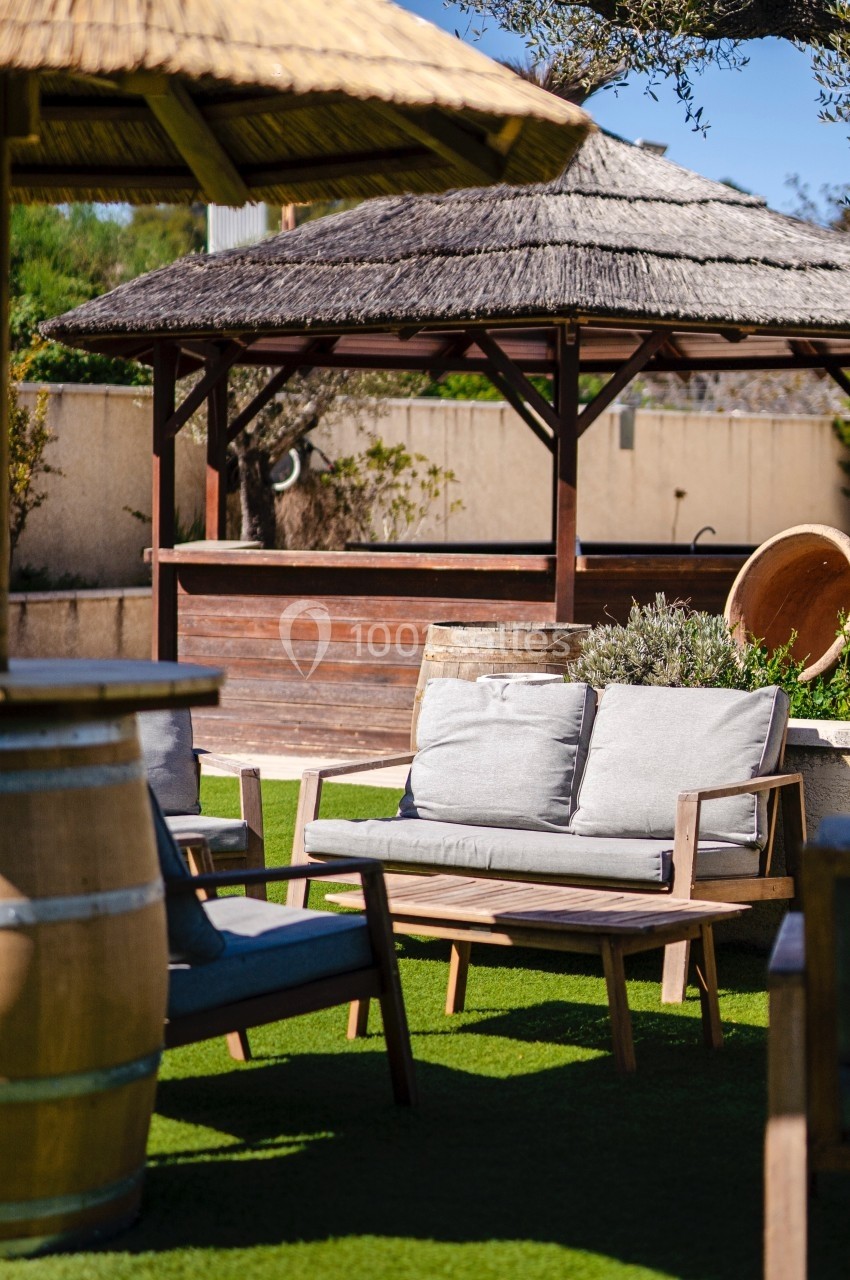 Mobilier de jardin en bois avec coussins gris, installé sur une pelouse artificielle près de pergolas en paille.