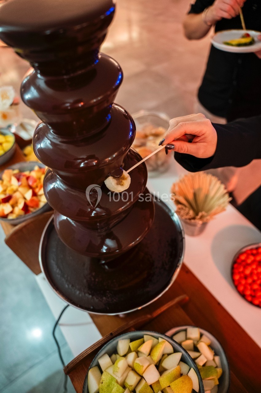 Fontaine de chocolat avec une personne trempant un morceau de fruit sur une brochette, entourée de fruits variés.