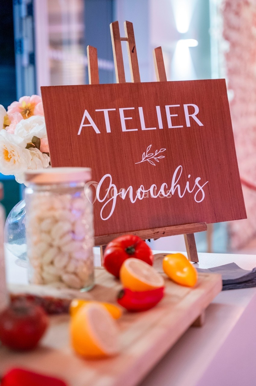 Panneau en bois indiquant ’Atelier Gnocchis’ avec des légumes et un bocal de gnocchis en premier plan.