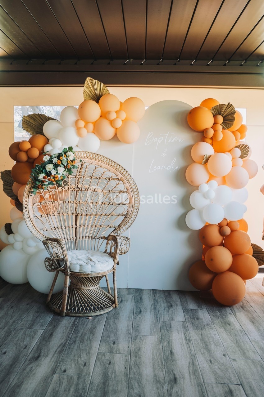 Fauteuil en rotin devant une arche décorée de ballons orange et blancs, dans un espace intérieur avec sol en bois.