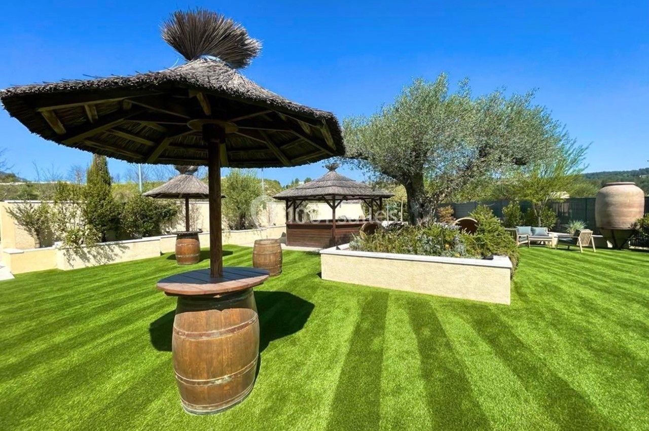 Espace extérieur avec pelouse artificielle, parasols en chaume, tonneaux en bois et végétation méditerranéenne.
