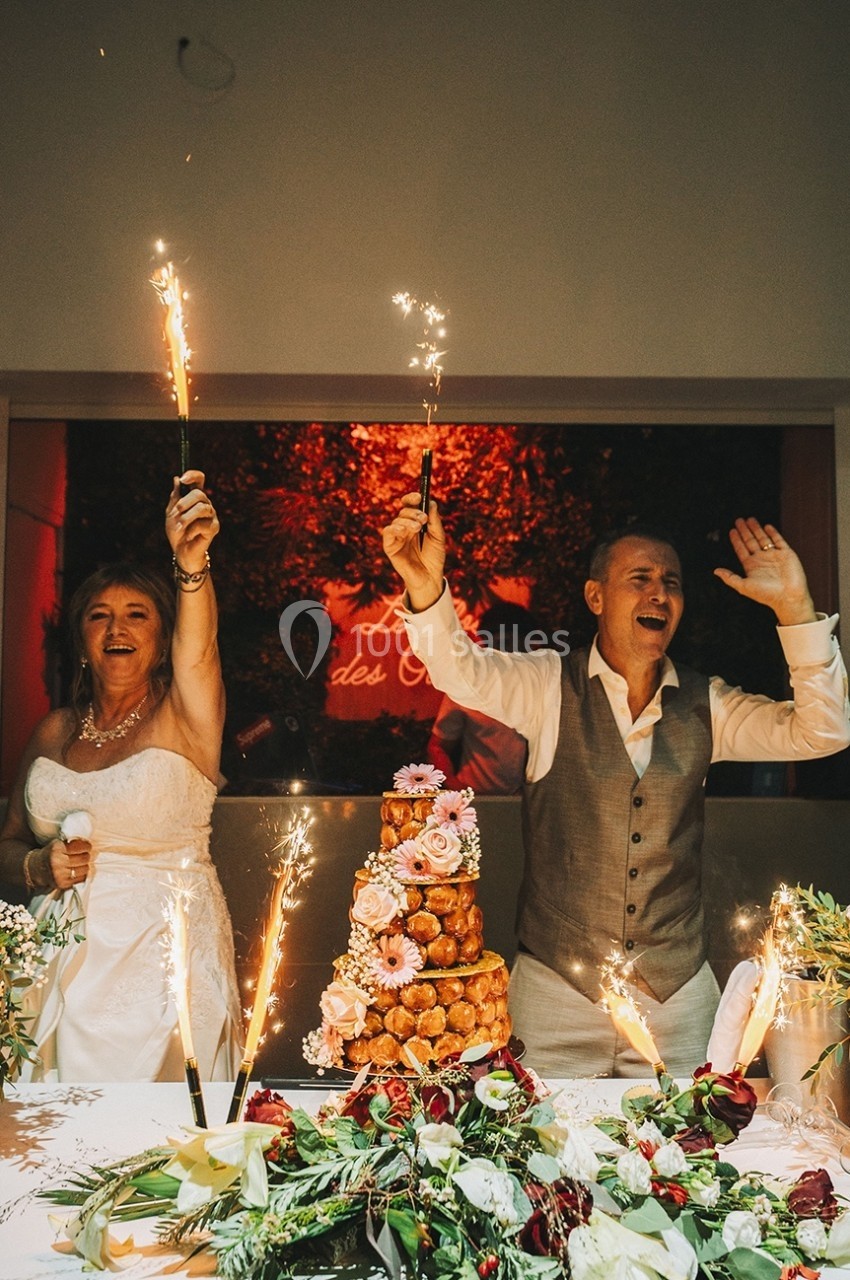 Un couple en tenue de mariage célèbre avec des cierges magiques devant une pièce montée décorée de fleurs.