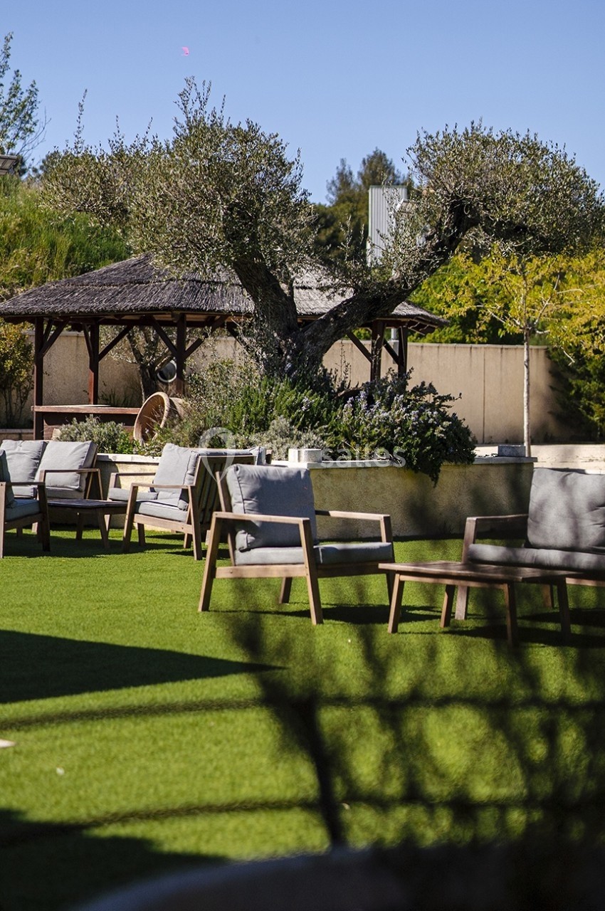 Chaises en bois avec coussins disposées sur une pelouse, près d'un olivier et d'une pergola en arrière-plan.