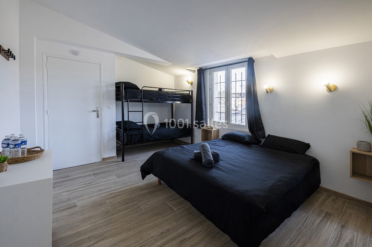 Chambre lumineuse avec un lit double, des lits superposés, sol en bois clair, murs blancs et fenêtre avec rideaux sombres.